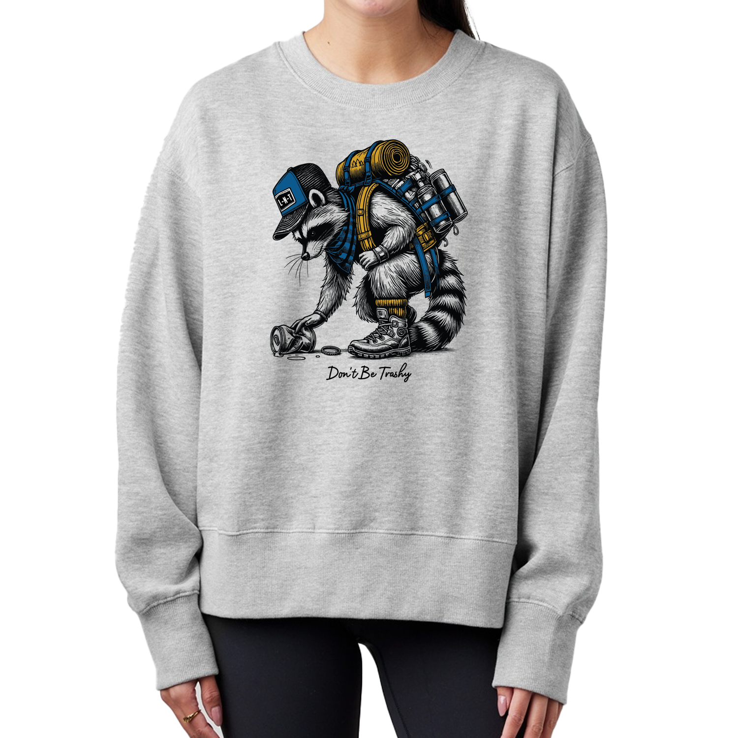Gray crewneck sweatshirt featuring an illustration of a hiking raccoon wearing a backpack and a cap, bending down to pick up a discarded can. The text "Don't Be Trashy" is written below the raccoon.