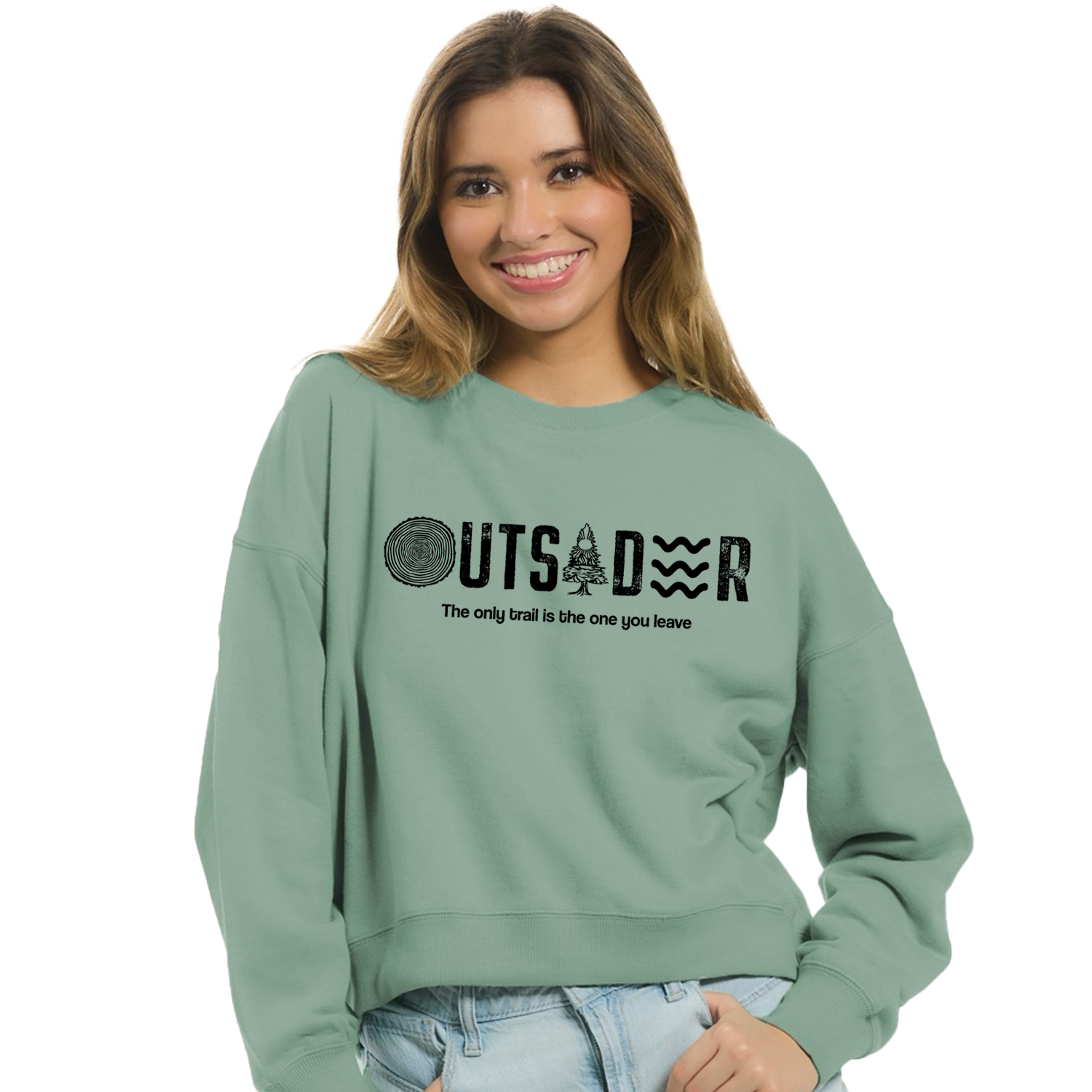 Woman wearing a cropped, sage green crewneck sweatshirt. The shirt features the word "OUTSIDER" in black, with letters stylized as a tree ring, a pine tree, and water/waves. Below the graphic, the tagline reads, "The only trail is the one you leave."
