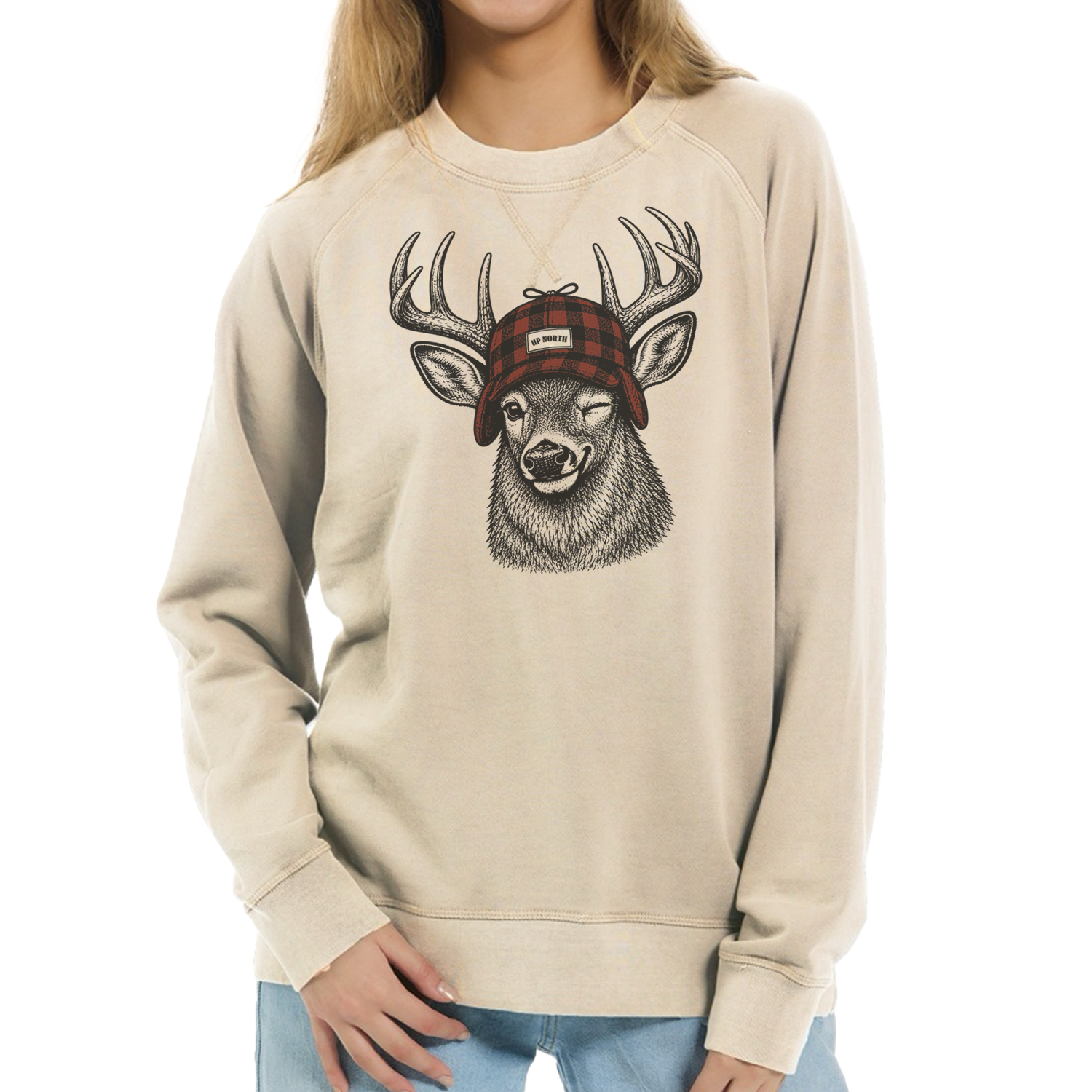 A person wearing a beige crewneck sweatshirt with a graphic of a deer head wearing a red plaid hat, paired with light blue jeans against a plain white background.